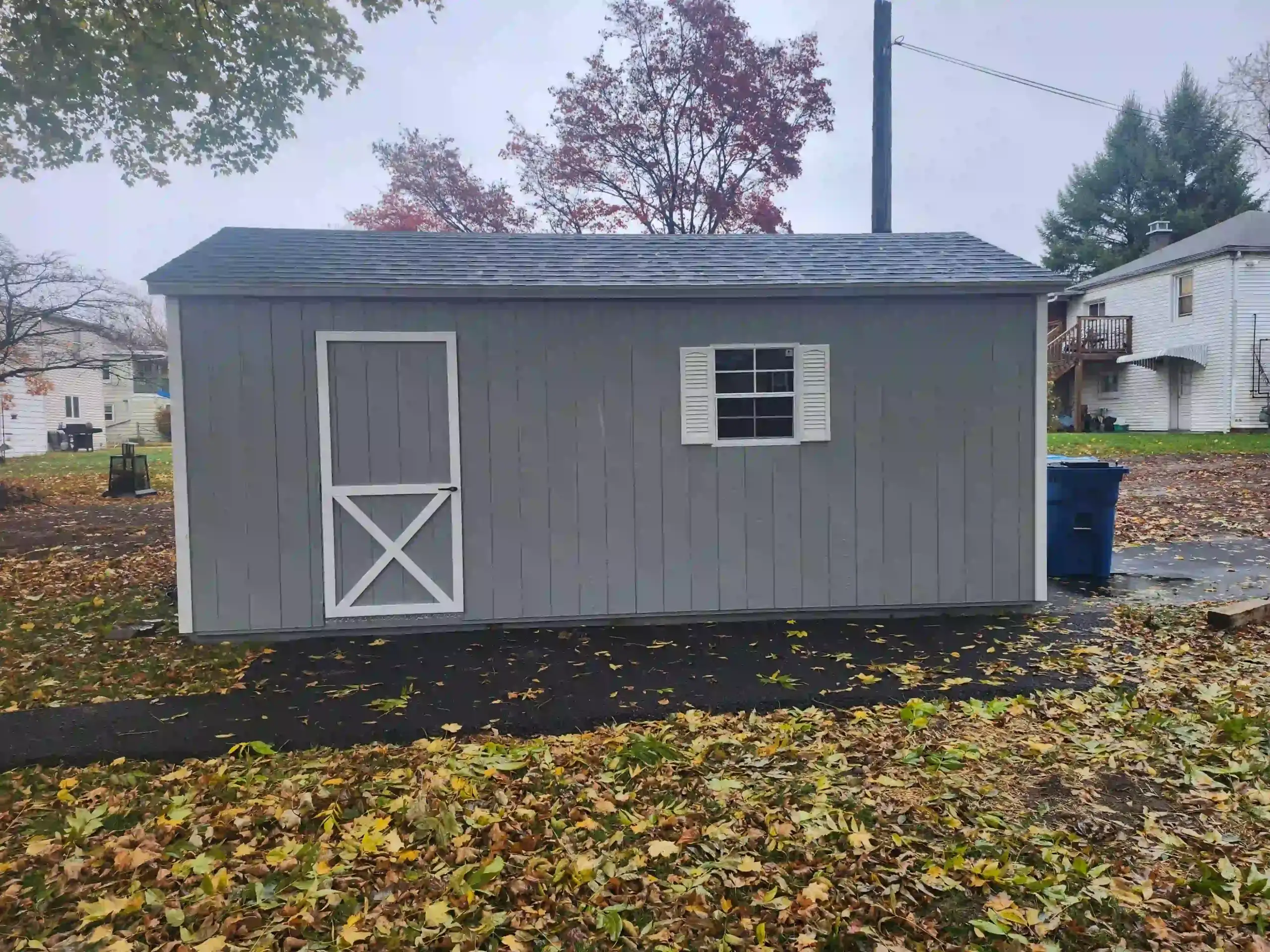 A gray single-car portable garage installed in a backyard, showing the convenient side entry door and window with shutters.
