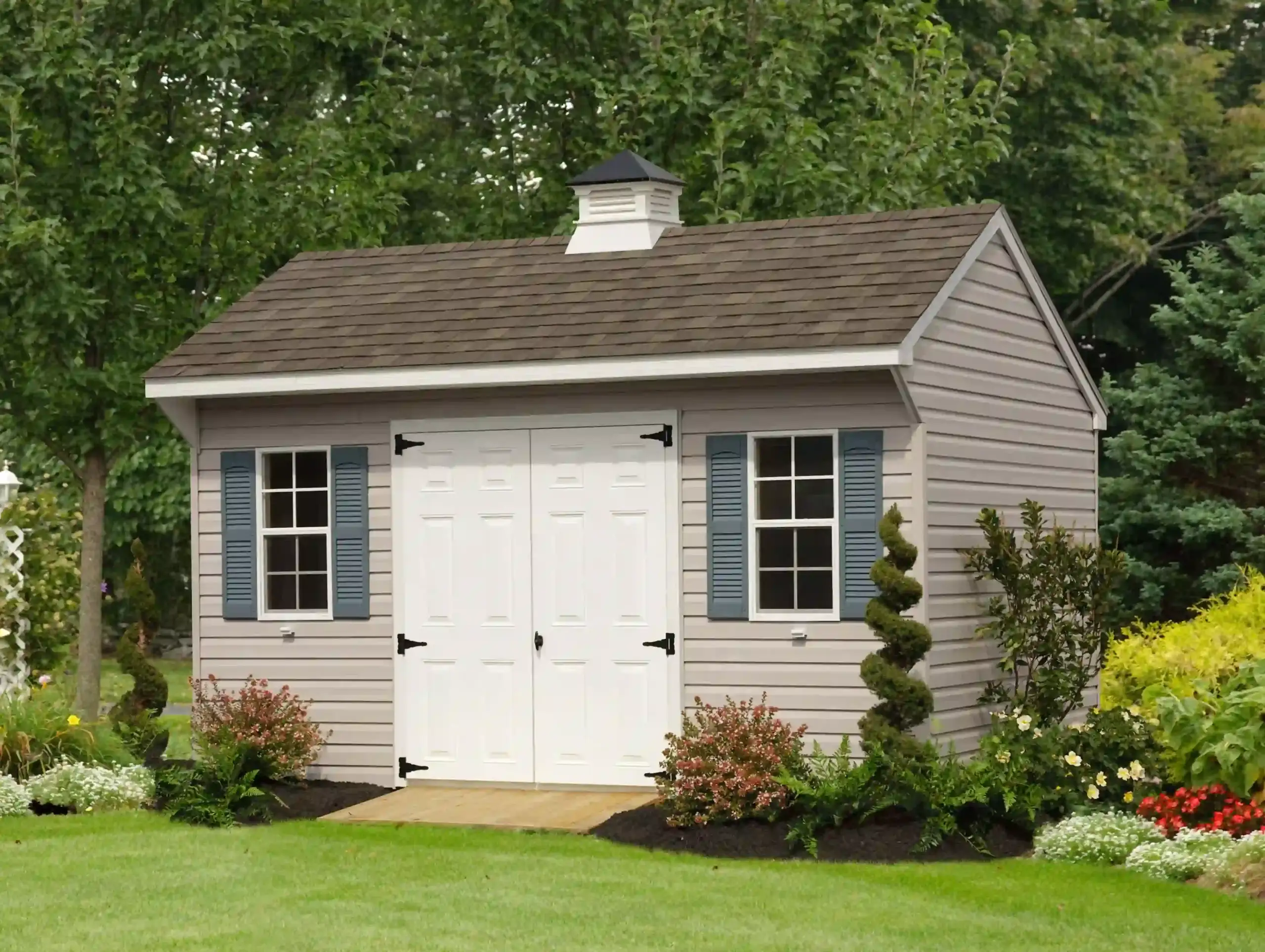 A classic vinyl A-frame shed with blue shutters and a decorative cupola, shown in a landscaped backyard setting, from Mill Run Storage.