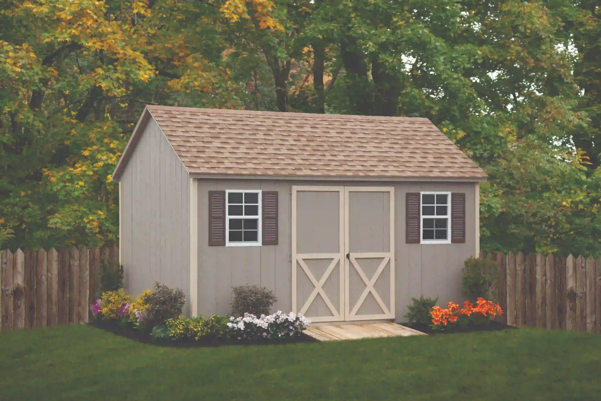 A tan wood garden shed with brown crossbuck doors and matching shutters, placed neatly in a backyard with a wooden fence.