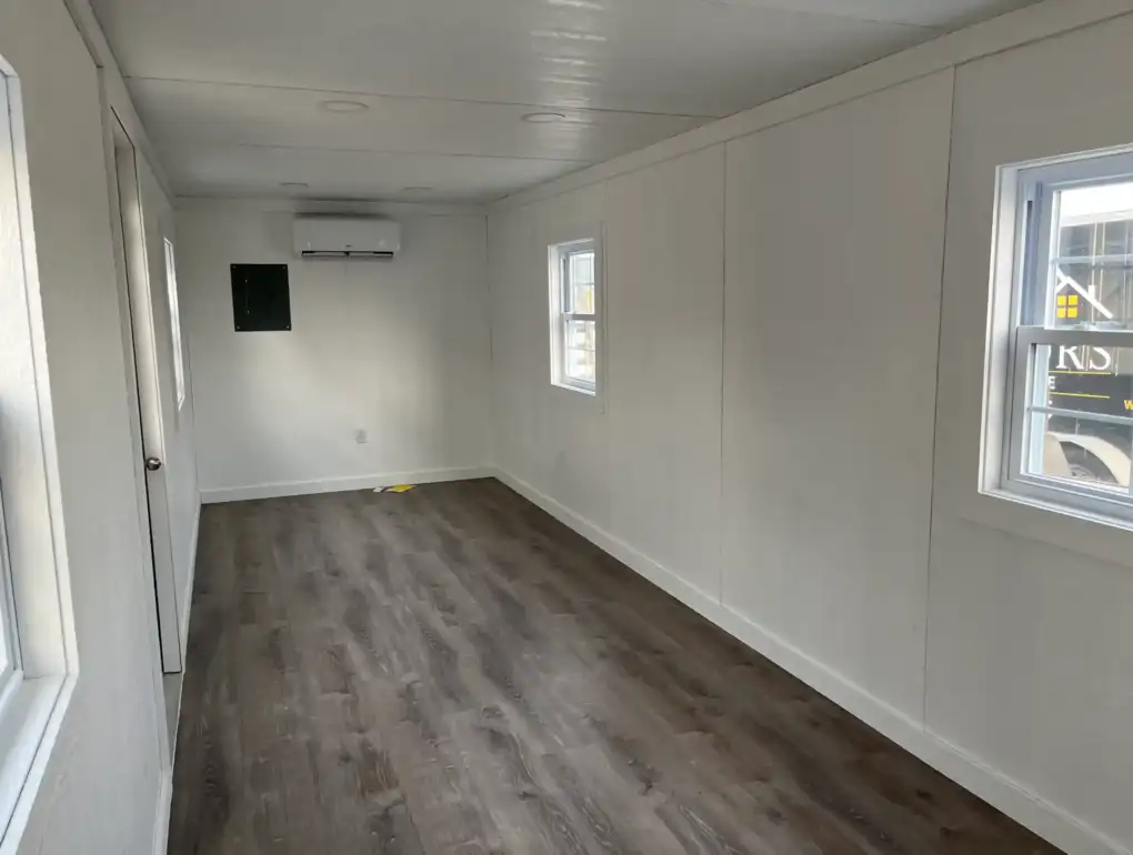 A finished portable office interior with white walls, wood-look flooring, an electrical panel, and a mini-split for heating and air conditioning.