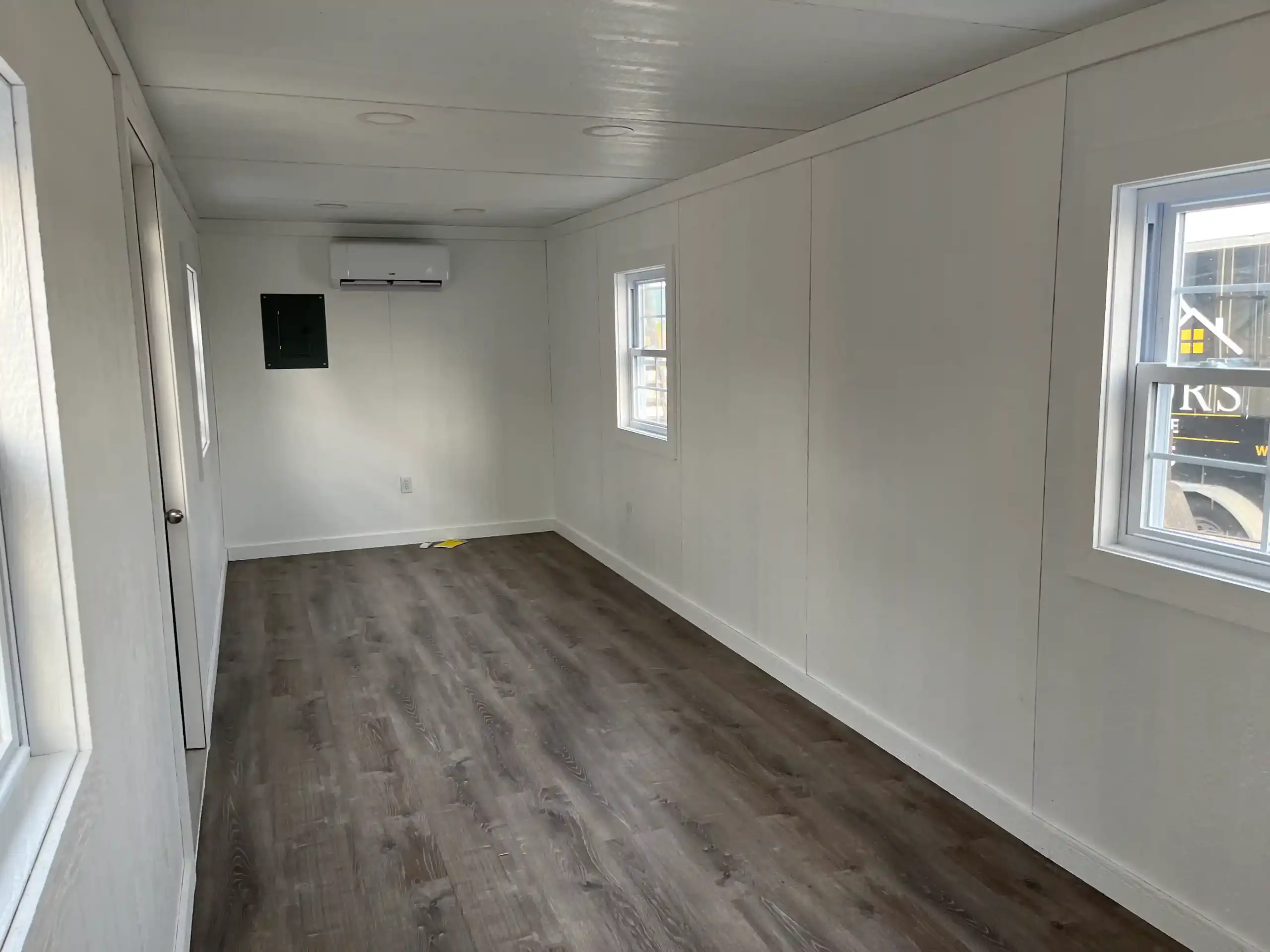 A finished portable office interior with white walls, wood-look flooring, an electrical panel, and a mini-split for heating and air conditioning.